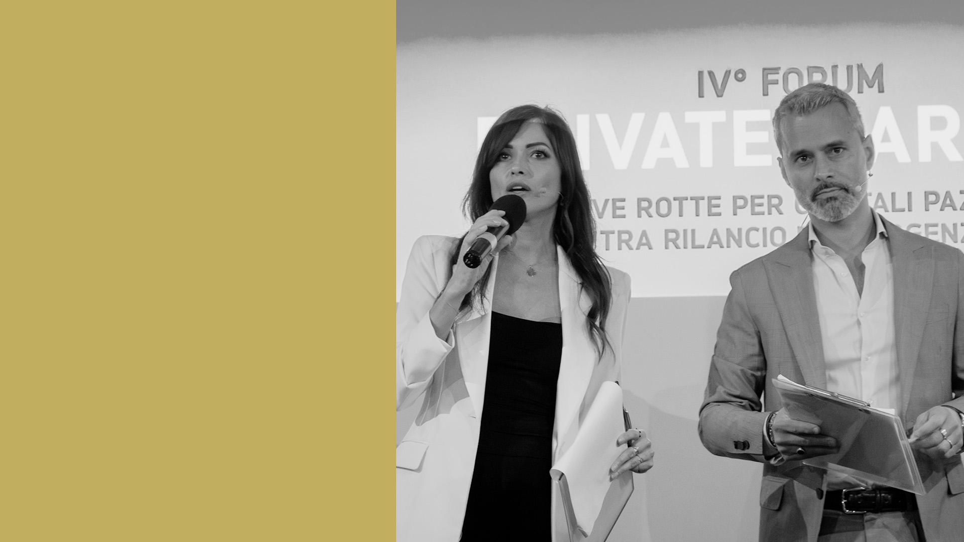 Una donna e un uomo in piedi su un palco, entrambi con in mano un microfono. La donna indossa un blazer bianco e l'uomo un completo chiaro. Stanno parlando a un evento con uno striscione sullo sfondo. Il lato sinistro dell'immagine ha un colore giallo-marrone uniforme.