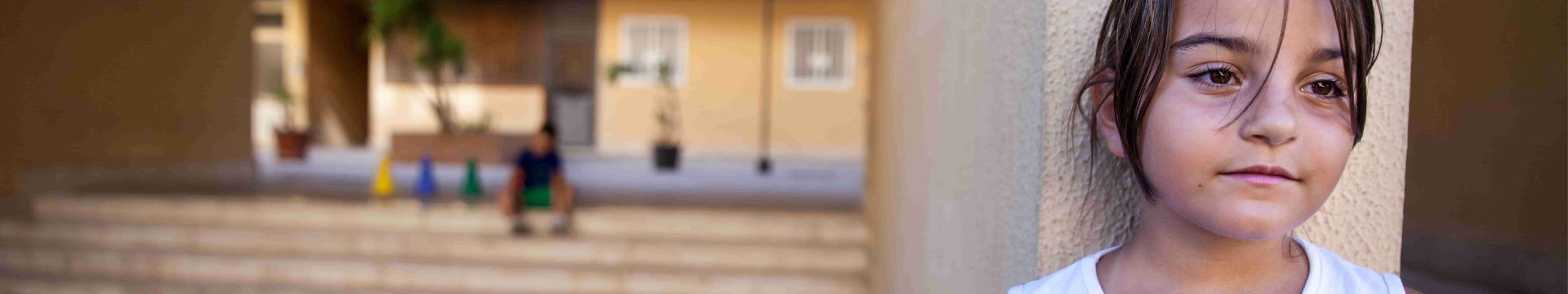Una giovane ragazza con un'espressione neutra è appoggiata a un muro, guardando in lontananza. Lo sfondo è sfocato, con una persona seduta sui gradini di fronte a un edificio.