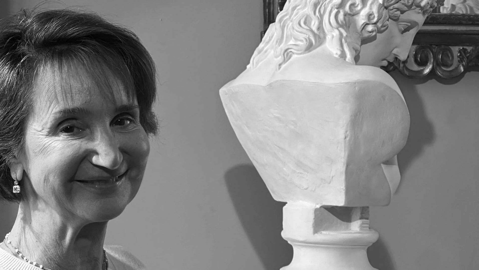 In questa fotografia in bianco e nero, una donna sorridente è in piedi accanto a un busto in marmo bianco raffigurante una figura dai capelli ricci, appoggiata contro una parete semplice, che riflette l'eleganza senza tempo delle collezioni italiane di Bassetti Christie.