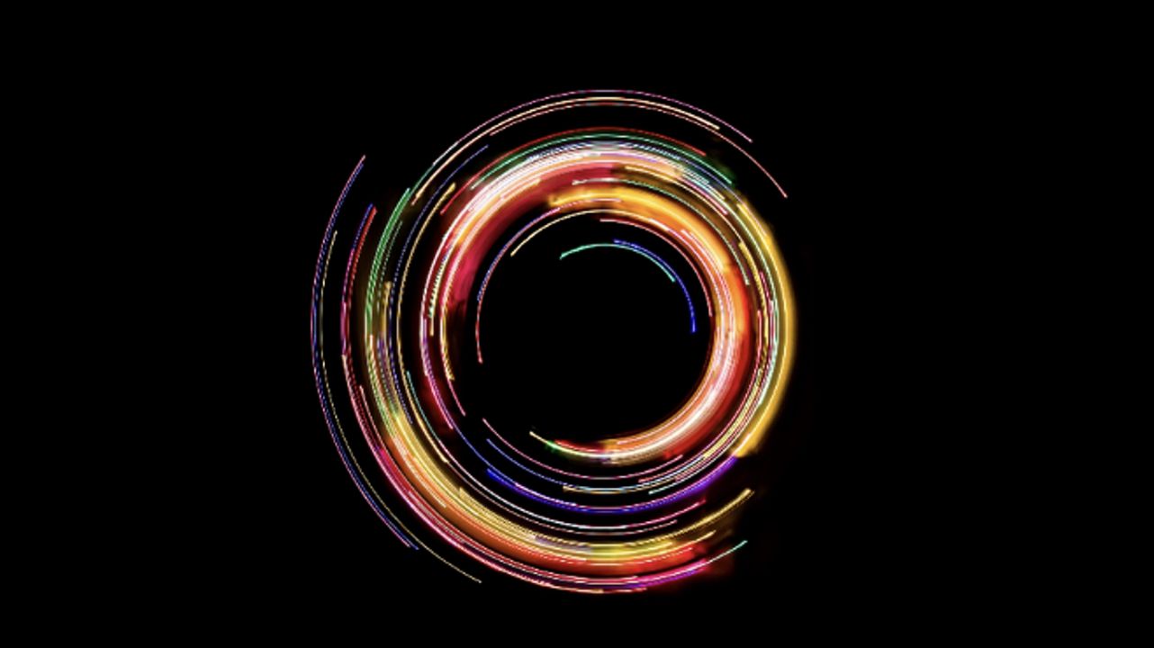 Un dipinto di luce circolare e dinamico su uno sfondo nero. L'immagine presenta scie luminose e vorticose di luci rosse, gialle e arancioni con accenni di verde e blu, creando un motivo a spirale vibrante.
