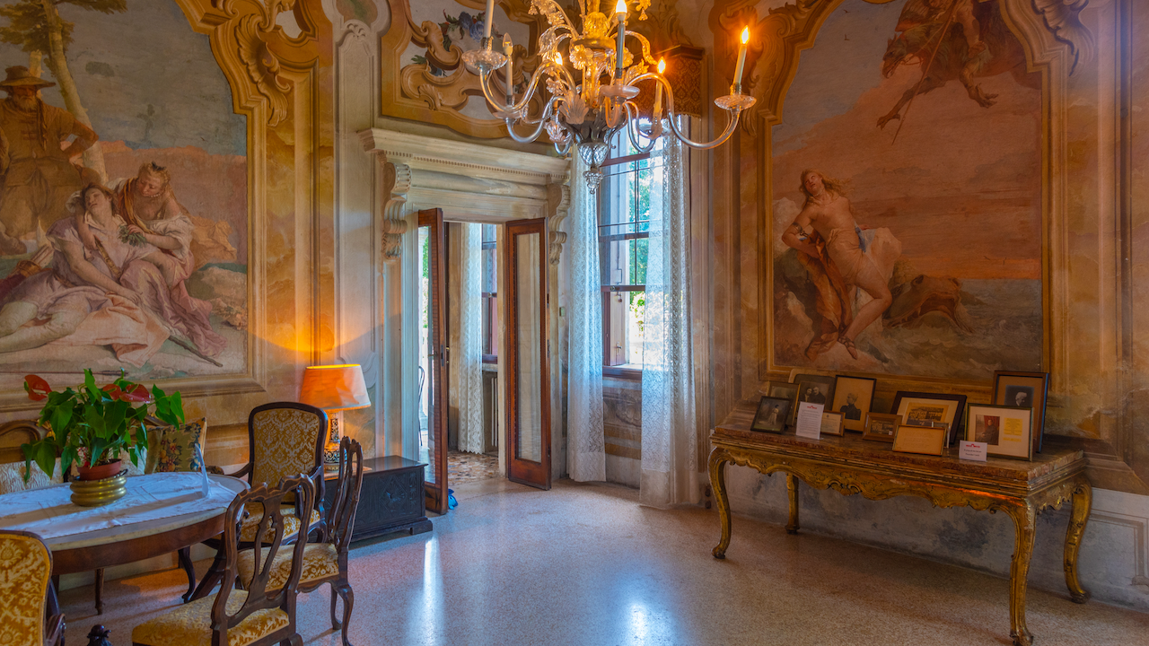 Elegante camera con pareti affrescate ornate con scene mitologiche. Un lampadario pende dal soffitto e un tavolo in legno con sedie si trova sulla sinistra. Una consolle con foto incorniciate si trova sulla destra e una porta aperta conduce all'esterno.