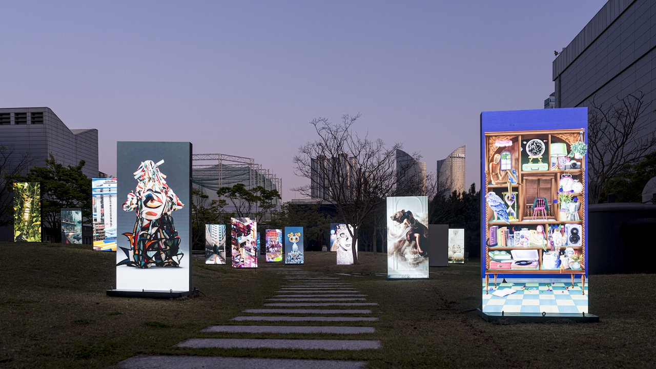 Una mostra notturna all'aperto presenta pannelli rettangolari illuminati che ospitano varie opere d'arte, tra cui dipinti, collage e arte digitale, disposti lungo un sentiero su un'area erbosa con edifici sullo sfondo.