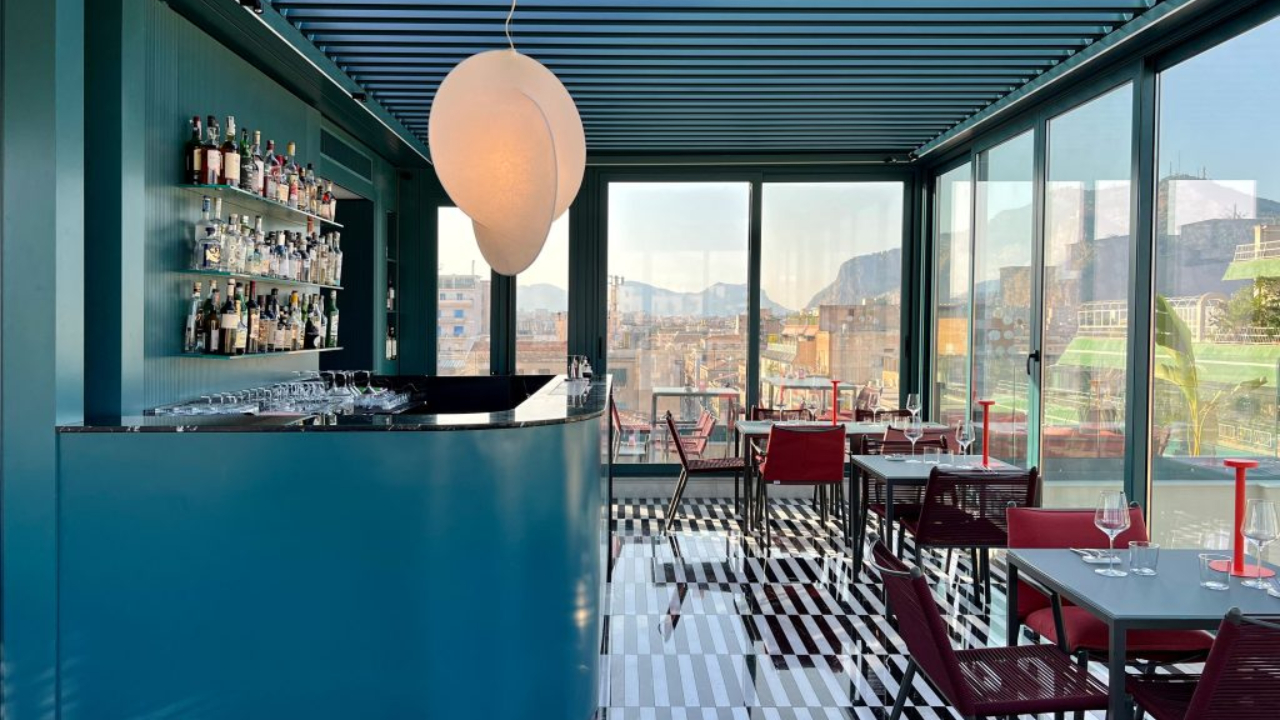 Moderno bar panoramico a Palermo con pareti color verde acqua, pavimento a righe bianche e nere, sedie rosse, tavoli in vetro e ampie finestre che offrono una vista panoramica sulla città e sulle montagne. Una lampada a sospensione bianca illumina il bancone curvo, ricco di bottiglie.
