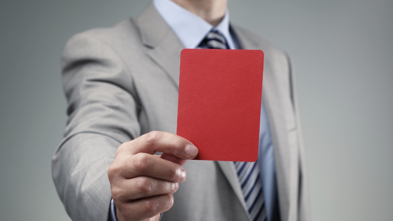 Una persona in abito grigio e cravatta a righe tiene un cartellino rosso verso la telecamera su uno sfondo grigio. L'attenzione è sulla mano e sul cartellino.