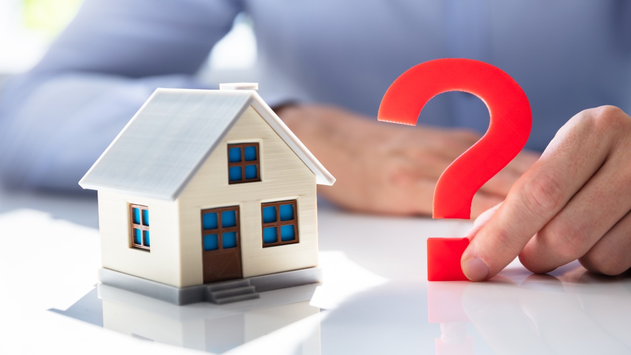Una persona tiene in mano un grande punto interrogativo rosso accanto a un piccolo modellino di casa su una superficie bianca, per simboleggiare l'incertezza nelle decisioni immobiliari e nel tipo di contratto da stipulare tra affitti brevi e canone concordato.