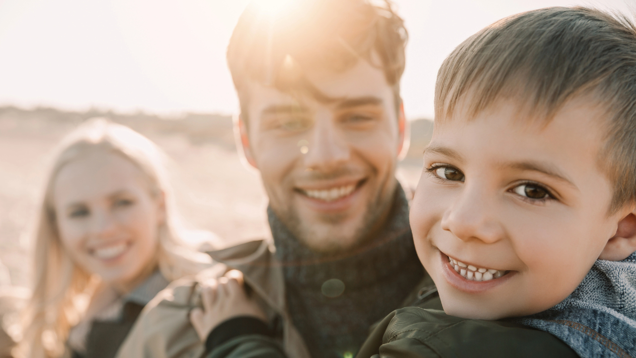 Un bambino sorridente in primo piano è tenuto in braccio da un uomo, con una donna in piedi accanto a loro. Sono all'aperto, di fronte alla telecamera, mentre la luce del sole crea un'atmosfera calda e luminosa: catturare questo momento è gratificante quanto ricevere un assegno familiare.