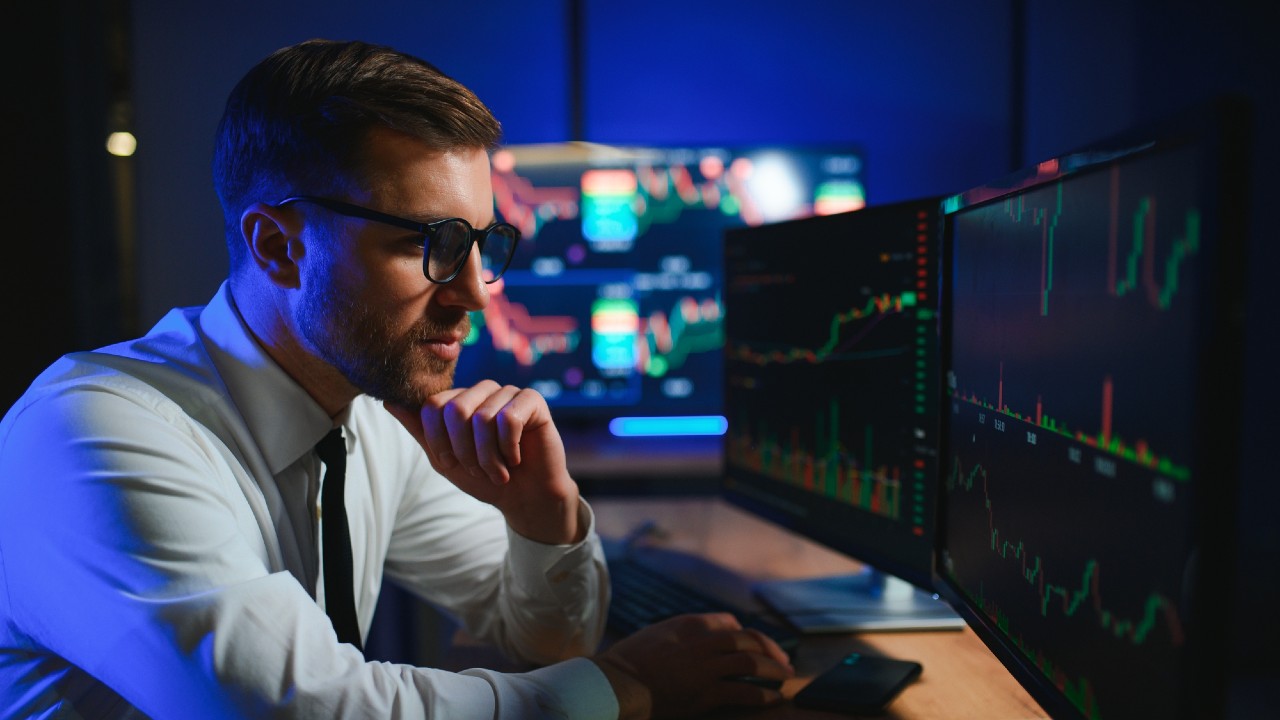 Un uomo in camicia bianca e cravatta, con gli occhiali, è seduto a una scrivania e sta analizzando grafici e diagrammi azionari visualizzati su più monitor di computer in un ufficio scarsamente illuminato.