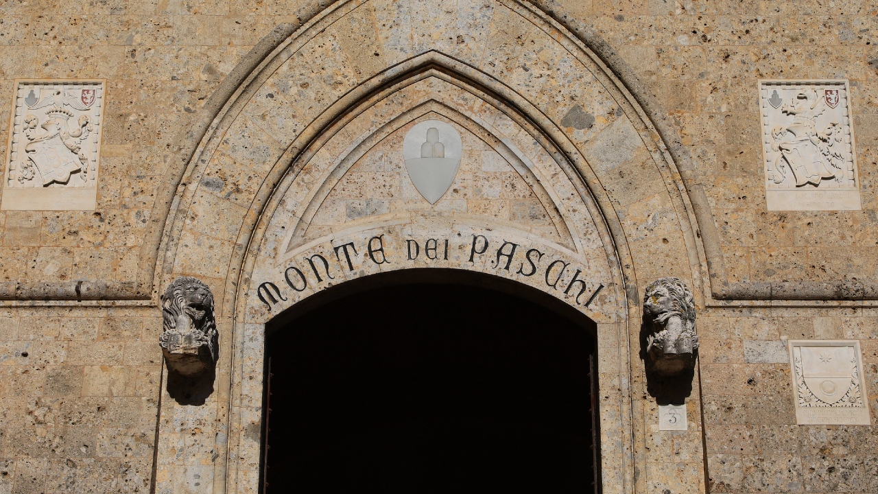 Ingresso ad arco in pietra con la scritta "Monte dei Paschi", fiancheggiato da due leoni scolpiti. Un accenno all'eleganza del MPS è dato dallo stemma triangolare in pietra sovrastante, con intricati rilievi decorativi che ornano il muro circostante.