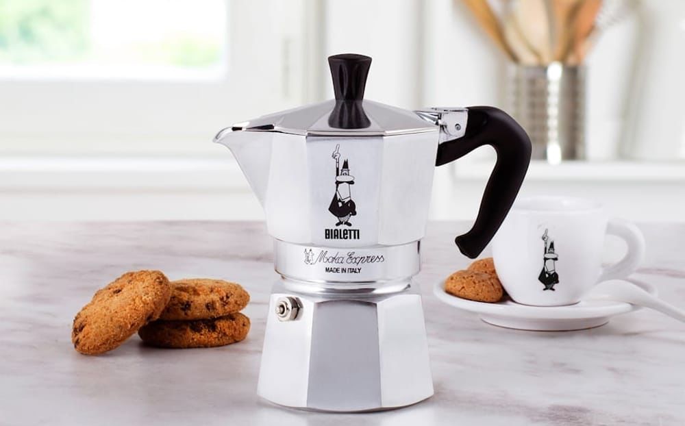 Una caffettiera Bialetti Moka Express argentata da piano cottura è appoggiata sul bancone della cucina, accanto a una tazza di caffè con lo stesso logo e a una piccola pila di biscotti al cioccolato. Sullo sfondo si vedono utensili da cucina sfocati.