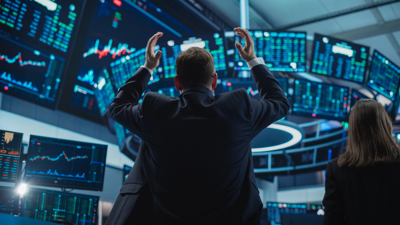 Una persona in giacca e cravatta con le braccia alzate è di fronte a più grandi schermi che mostrano dati e grafici del mercato azionario, catturando l'energia frenetica di un ambiente di trading di borse. Nelle vicinanze, un altro individuo è pronto nella sala di trading finanziario.