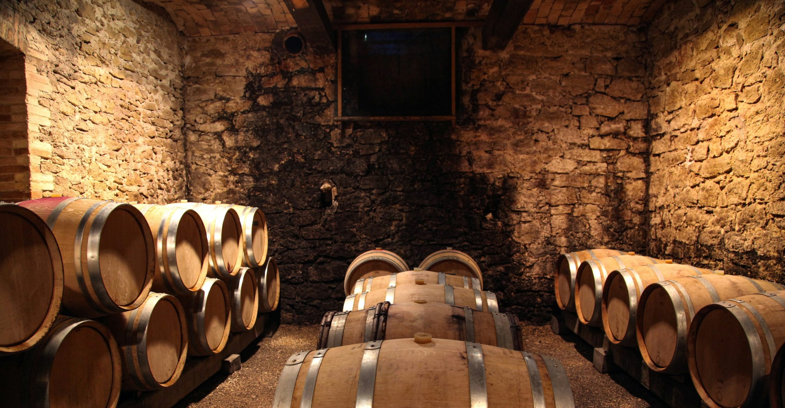 Una cantina scarsamente illuminata con pareti in pietra e soffitto in mattoni evoca il fascino senza tempo spesso notato nelle notizie sul vino. Diverse botti di legno sono accatastate contro le pareti, aggiungendo all'atmosfera rustica che sottolinea l'antica tradizione della vinificazione.