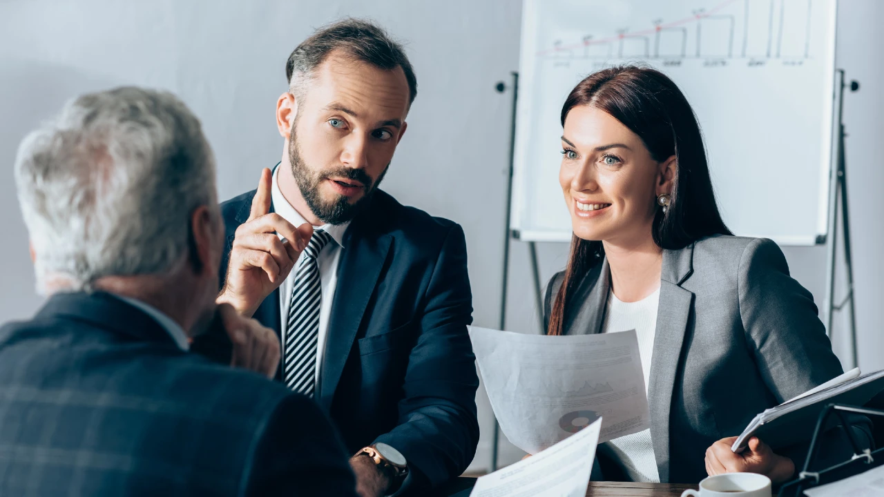 In una riunione, tre professionisti discutono di strategie. Un uomo barbuto in giacca e cravatta gesticola con entusiasmo su come investire 100 euro, mentre una donna in blazer grigio regge documenti e sorride. Un uomo anziano con i capelli grigi ascolta attentamente, con i grafici visibili sulla lavagna alle loro spalle.