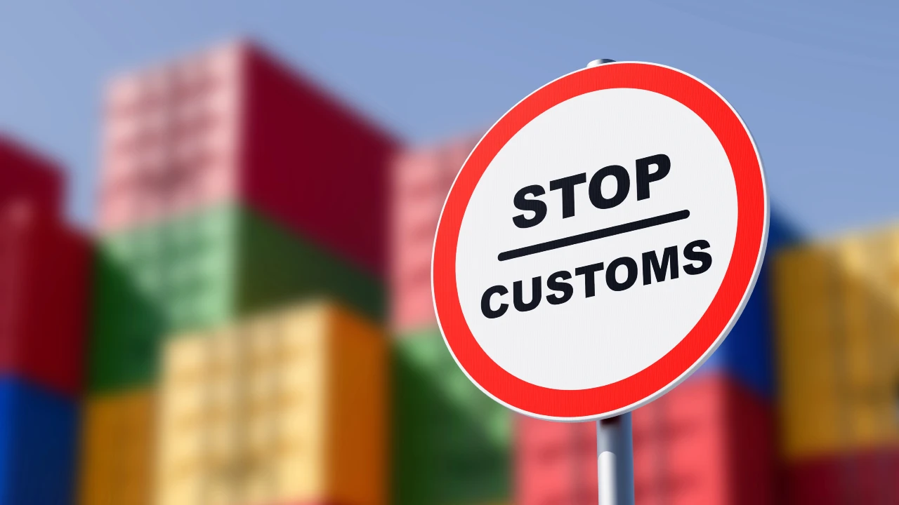 Un cartello circolare con la scritta "STOP DOGANA" è ubicato di fronte a container colorati e impilati, con un cielo azzurro e limpido sullo sfondo. Il riferimento va a dazi ambientali e alla norma Dpam