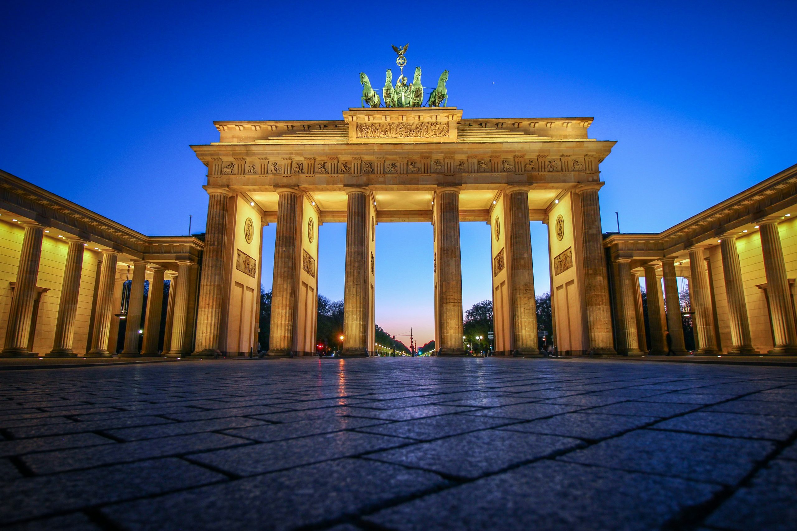 La Porta di Brandeburgo a Berlino, in Germania, è illuminata al tramonto sullo sfondo di un cielo azzurro terso, se osservata da un'angolazione bassa sulla piazza acciottolata antistante.