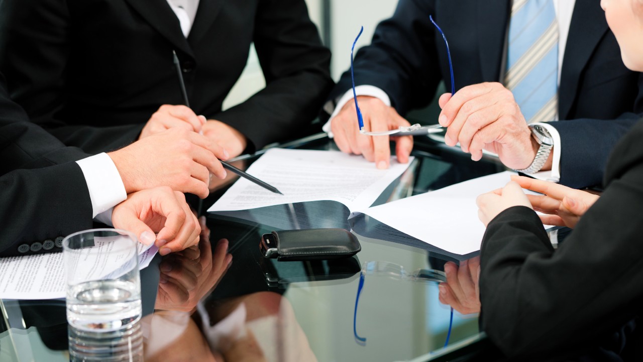 Diverse persone in abiti da lavoro sono sedute a un tavolo di vetro e discutono di alcuni documenti. L a foto vuole evidenziare il tema della collaborazione in un ambiente professionale incentrato sulle imprese familiari.