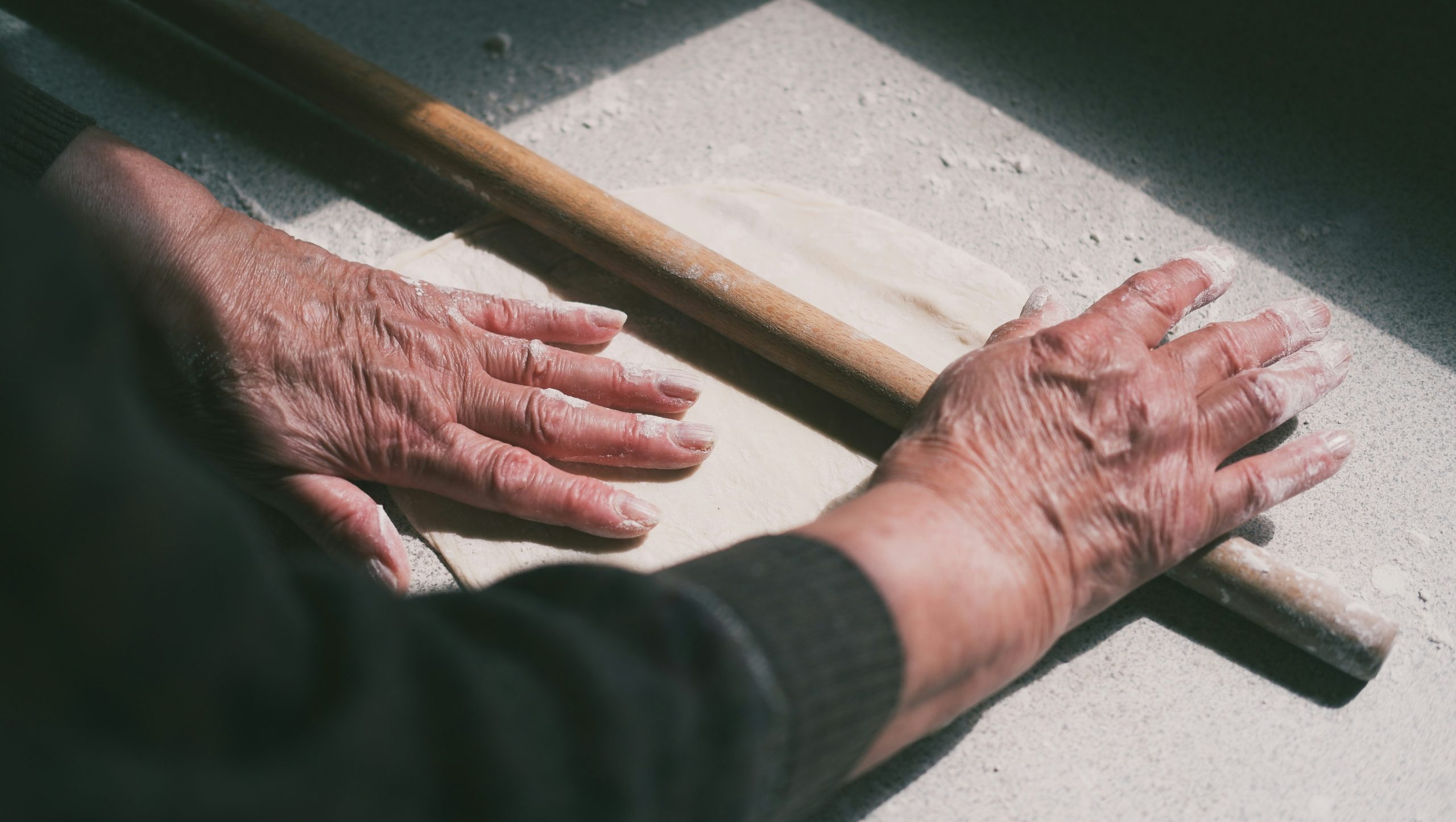 Una persona con le mani rugose stende l'impasto con un mattarello di legno su un tavolo infarinato, illuminato dalla luce naturale del sole.