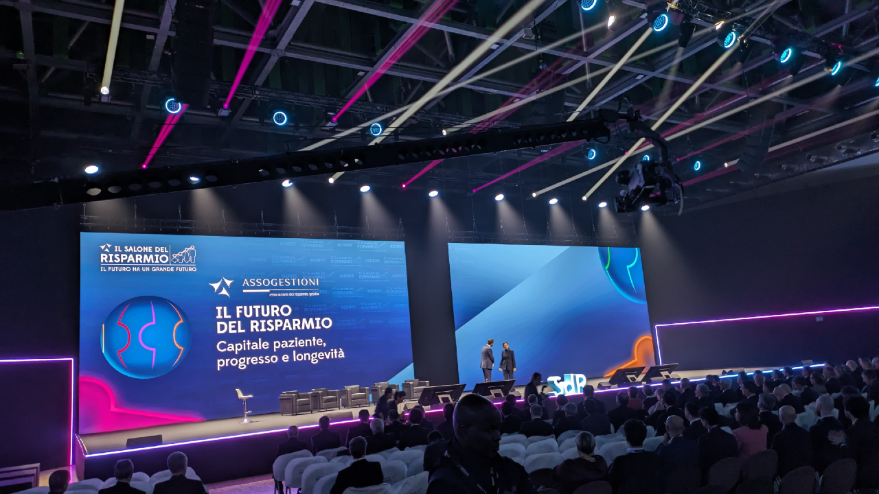 Un'ampia sala conferenze con un palco illuminato presenta "Il Futuro del Risparmio: Capitale paziente, progresso e longevità", mentre colori vivaci e schermi si fondono perfettamente, evocando un ambiente moderno e high-tech, che ricorda l'estetica elegante del design Gota.