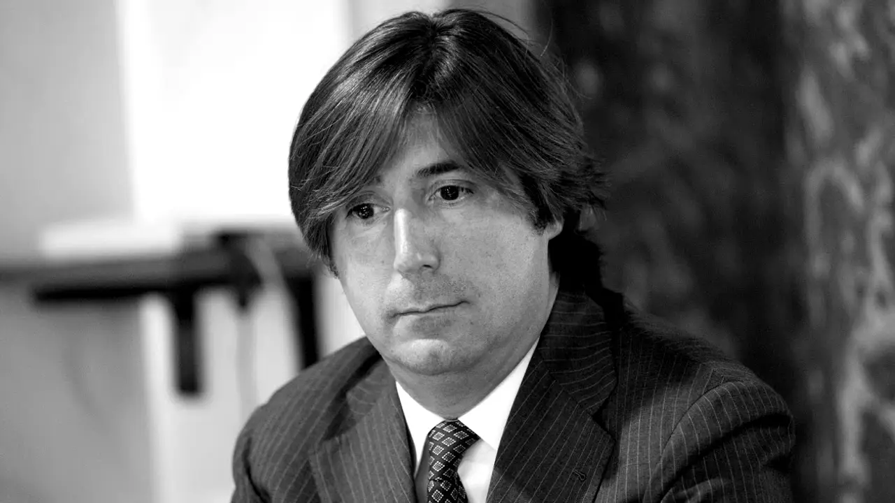 Un uomo in giacca e cravatta, con i capelli di media lunghezza, guarda leggermente di lato con un'espressione neutra. L'immagine è in bianco e nero.