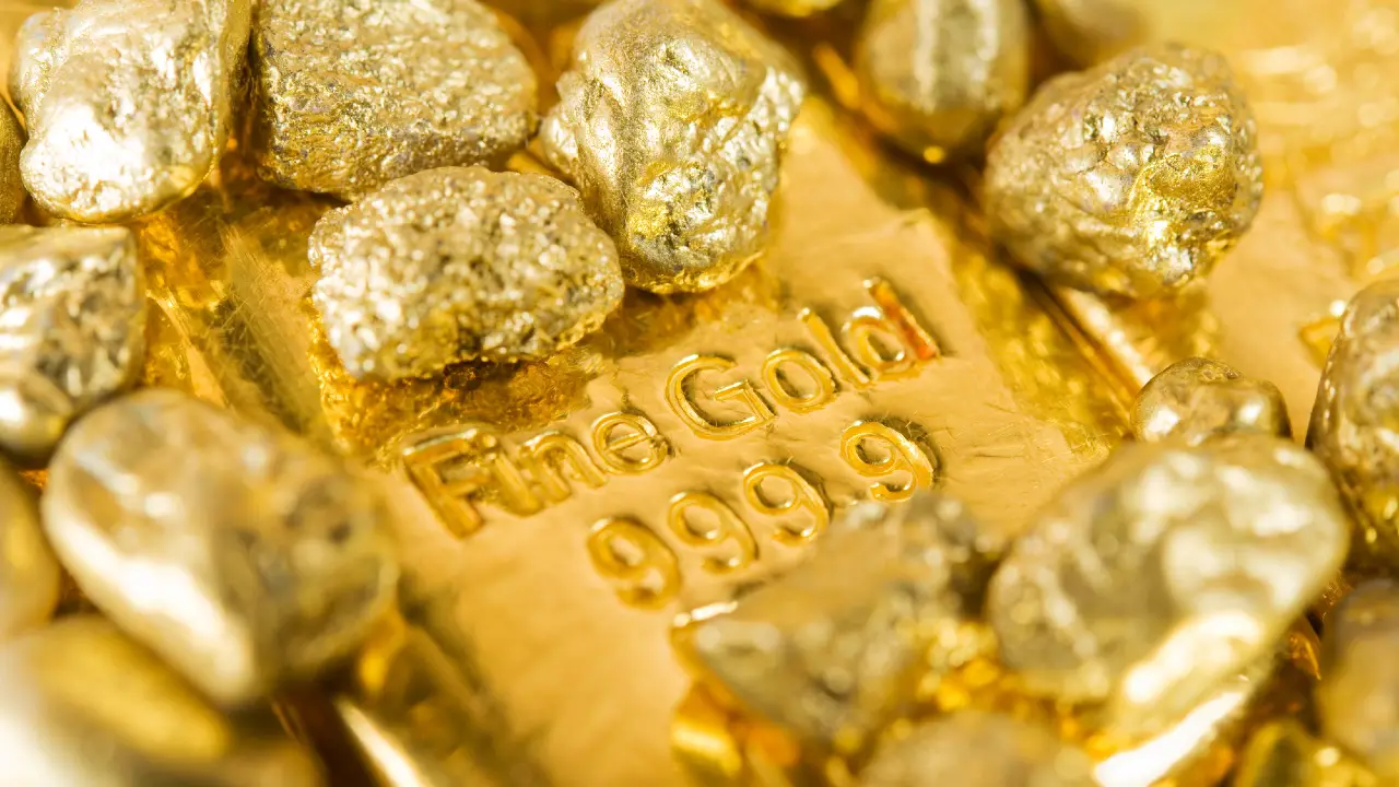 Primo piano di una barra d'oro con la scritta "Fine Gold 999.9" circondata da numerose piccole pepite d'oro, tutte che riflettono una luce intensa.