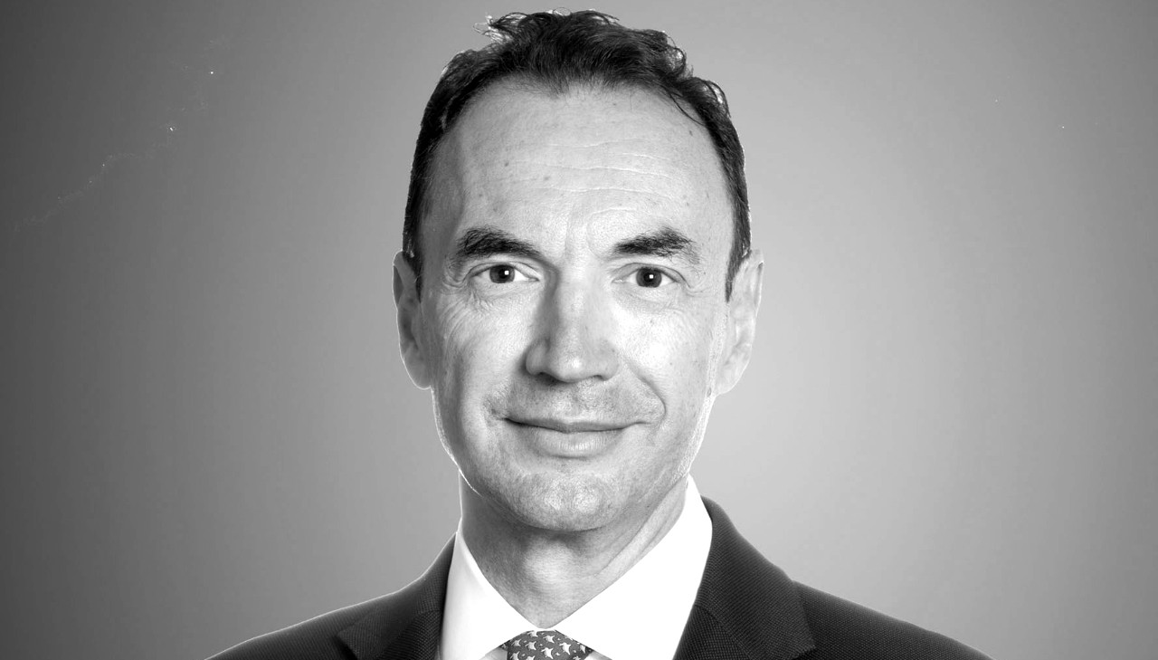 Un uomo vestito in modo professionale è ritratto in un ritratto in scala di grigi. Ha i capelli corti e scuri e indossa un abito con cravatta. È rivolto in avanti con un leggero sorriso su uno sfondo semplice.