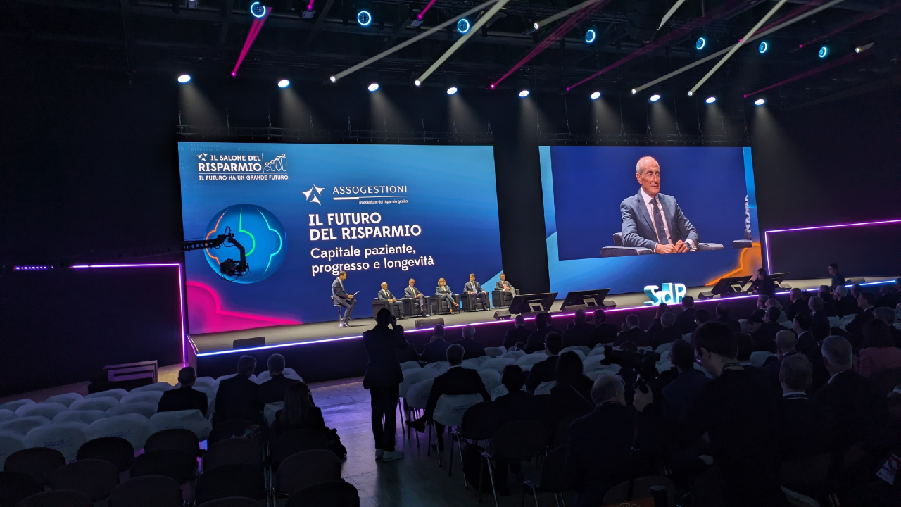 Un palco per conferenze con un gruppo di sei relatori seduti. Un grande schermo mostra il titolo dell'evento "Il Futuro del Risparmio". Il pubblico è seduto in file e l'illuminazione del palco crea un'atmosfera vibrante.