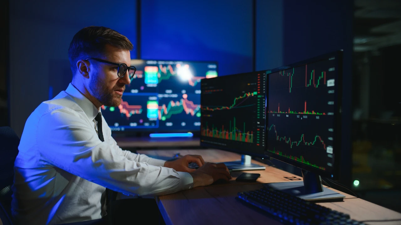Un uomo in camicia bianca e cravatta è seduto a una scrivania e lavora su diversi monitor su cui sono visualizzati grafici e diagrammi finanziari colorati in un ufficio scarsamente illuminato.