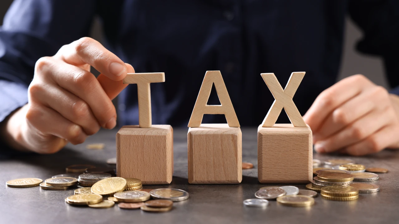 La mano di una persona dispone blocchi di legno con le lettere "T", "A" e "X" su un tavolo disseminato di monete, che simboleggiano concetti finanziari come tasse o trasferimenti.