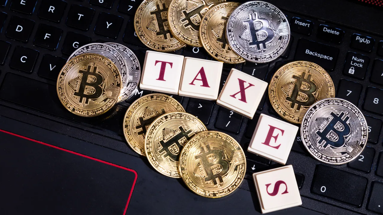 Monete Bitcoin d'oro e d'argento e altre criptovalute sparse sulla tastiera di un computer portatile con tessere dello Scrabble che compongono la scritta "tasse" in lettere rosse.
