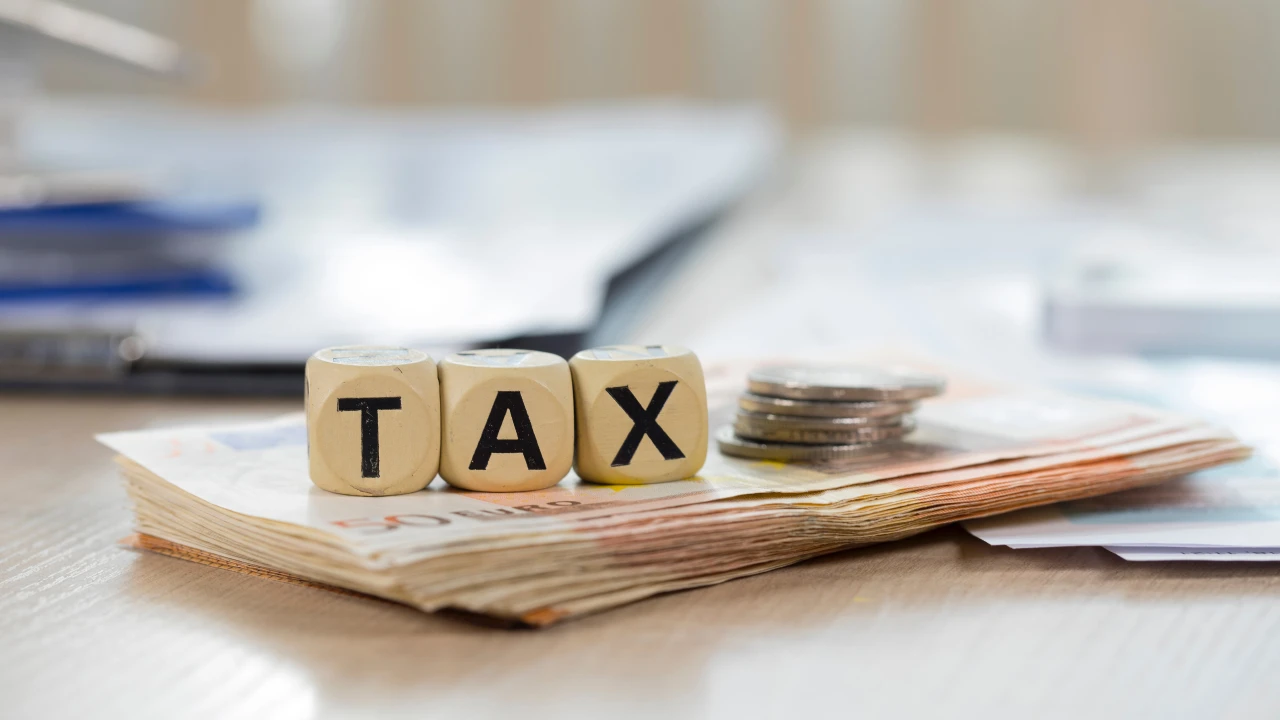 Dadi di legno con la scritta "TAX" sono appoggiati su una pila di banconote e monete, con documenti sfocati sullo sfondo. Il riferimento va alle novità fiscali sui trasferimenti di quote ai figli.