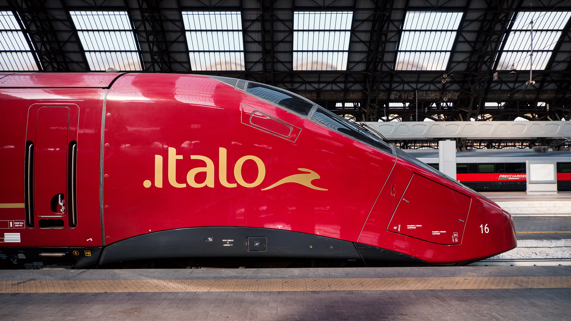 Un elegante treno rosso ad alta velocità Italo, perfetto per viaggiare, è parcheggiato su una banchina della stazione. Il design aerodinamico e il logo del treno sono in evidenza sotto il tetto in vetro e metallo ad arco. Un altro treno è visibile sullo sfondo, completando questa moderna scena di viaggio.