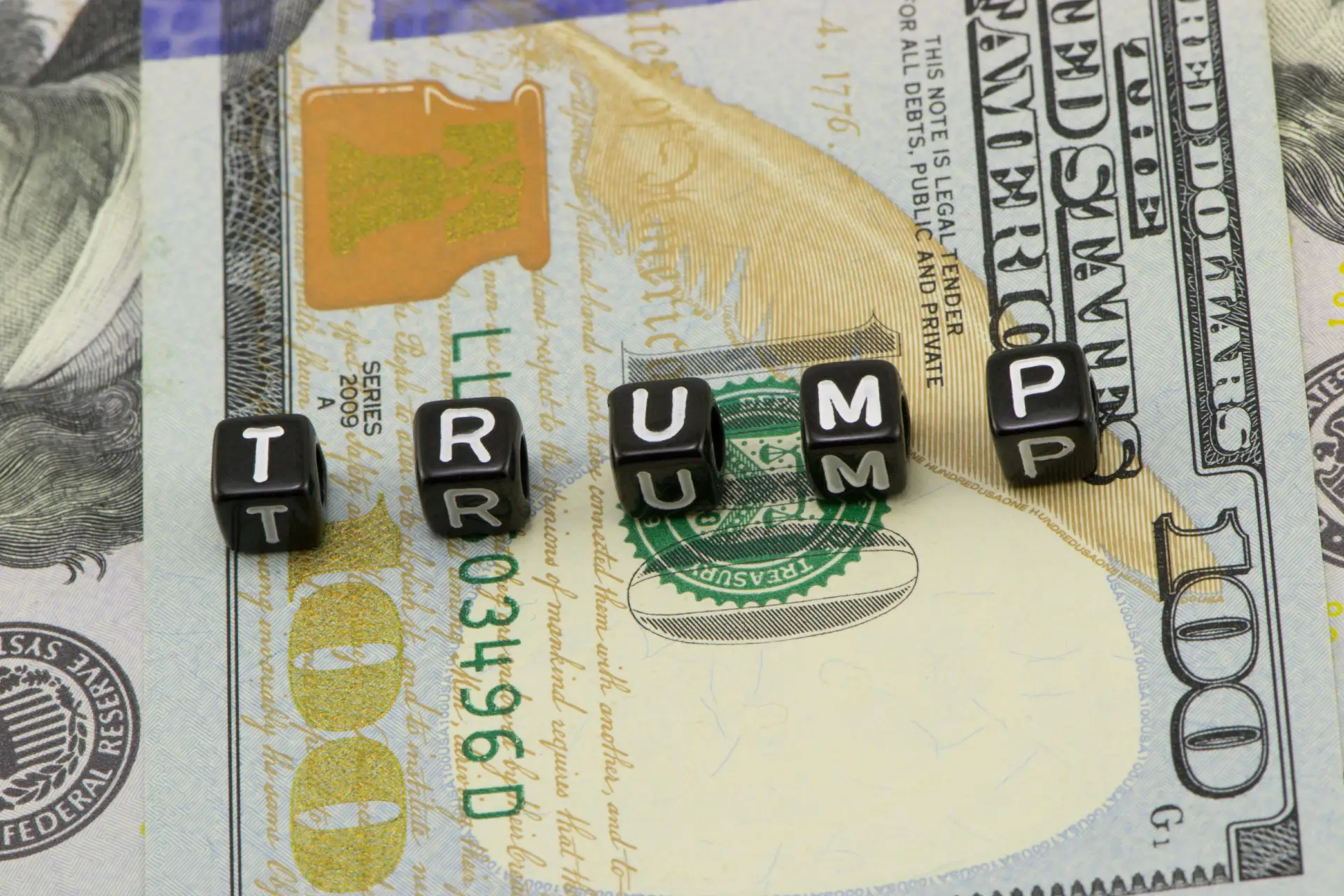 Su una banconota da 100 dollari americani sono presenti dei cubi con le lettere nere che compongono la scritta "TRUMP", con il ritratto e i dettagli della banconota visibili sullo sfondo.