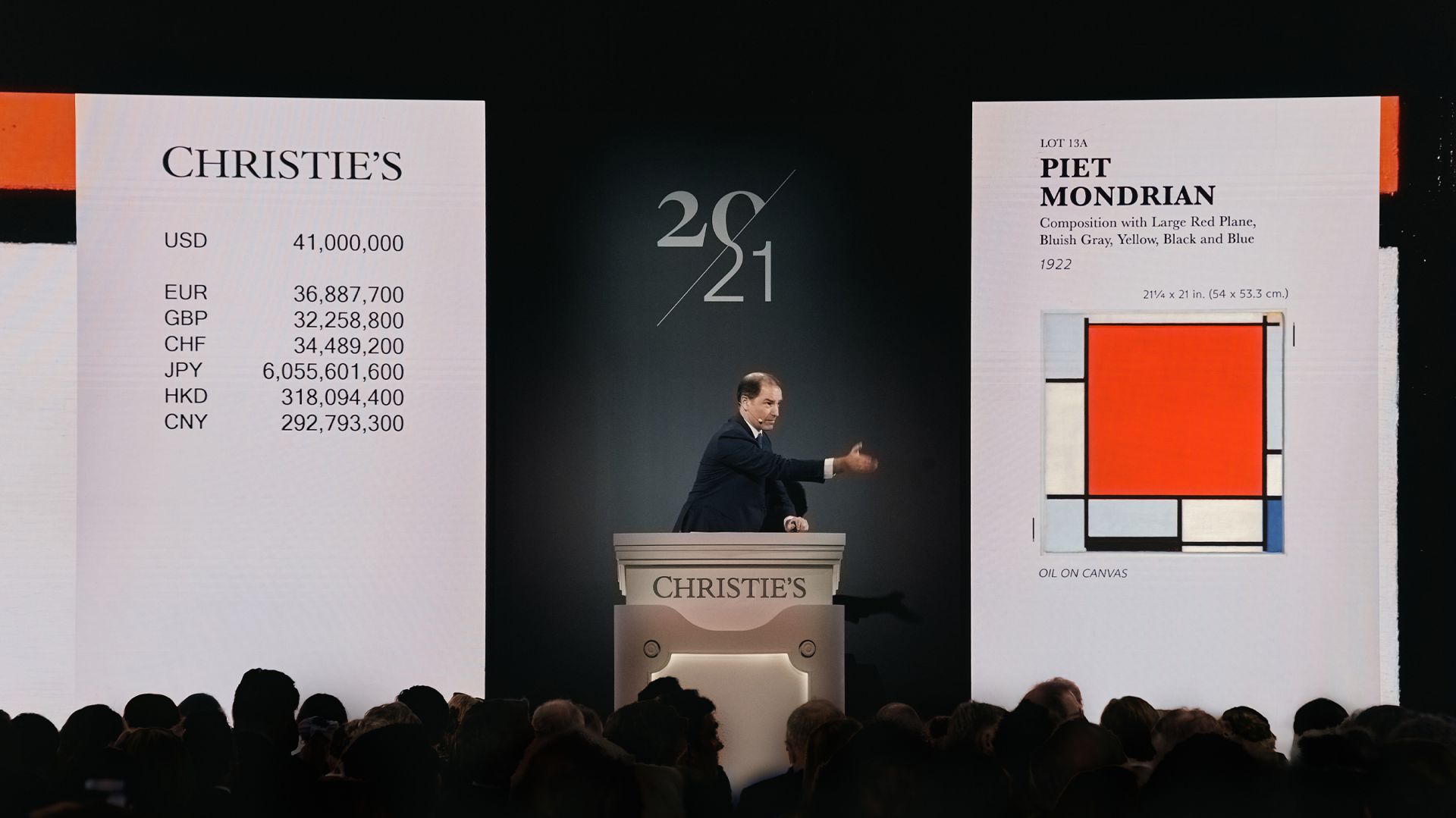 Un banditore d'asta di Christie's è in piedi su un podio, gesticolando durante un'asta d'arte. Degli schermi mostrano conversioni di valuta e dettagli di un dipinto di Piet Mondrian intitolato "Composizione con grande aereo rosso...".