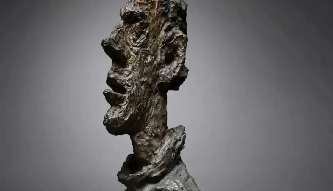 Un busto scolpito con una superficie ruvida e strutturata, raffigurante una testa umana sottile e allungata e un collo di profilo su uno sfondo grigio semplice.