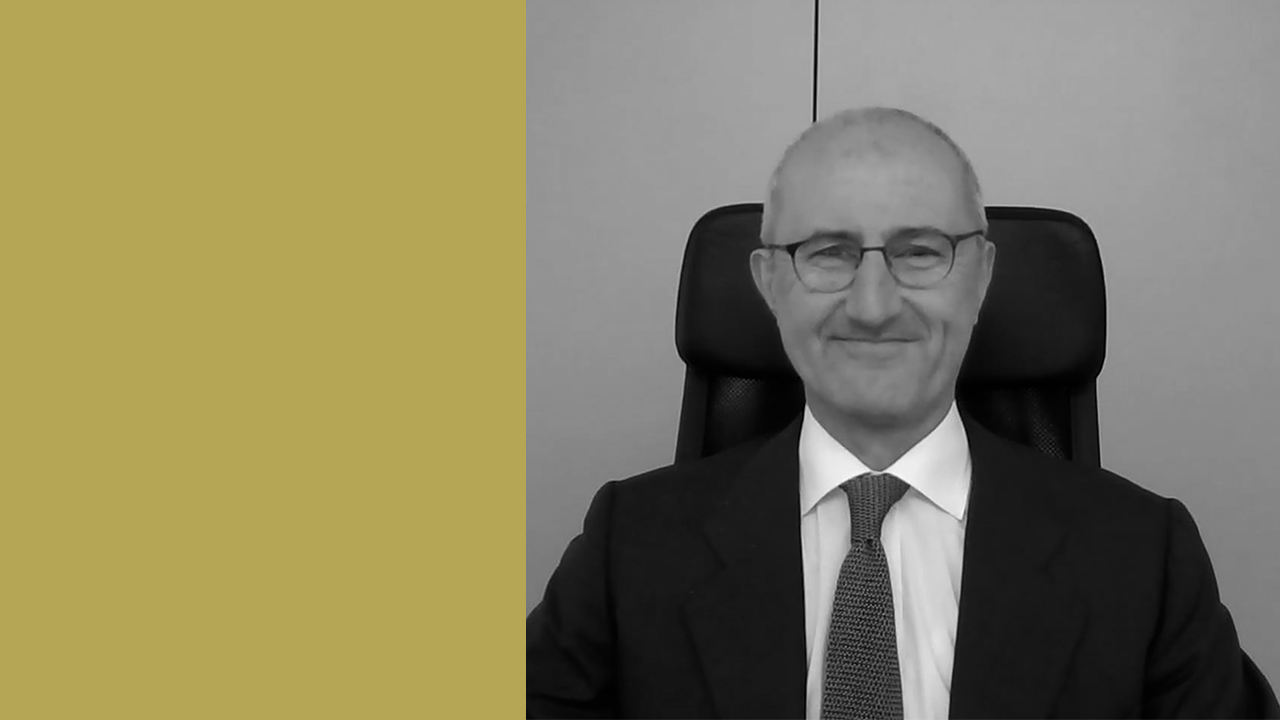 Un uomo in giacca e cravatta è seduto su una sedia da ufficio e sorride alla telecamera. L'immagine è in bianco e nero, con un grande rettangolo giallo senape sul lato sinistro.