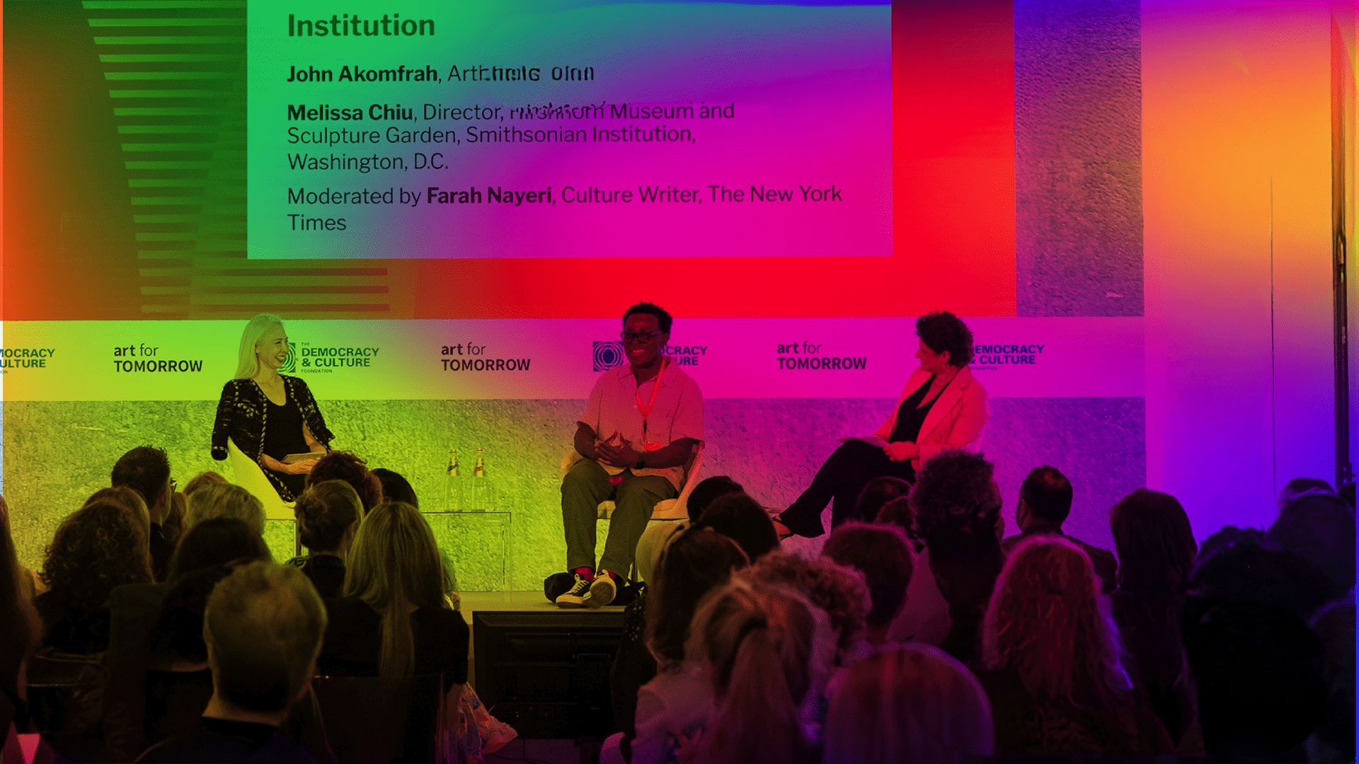 Tre relatori siedono sul palco davanti a uno sfondo colorato e illuminato durante la tavola rotonda di Art for Tomorrow, mentre il pubblico ascolta. Uno schermo in alto mostra i nomi dei relatori e i dettagli dell'evento.