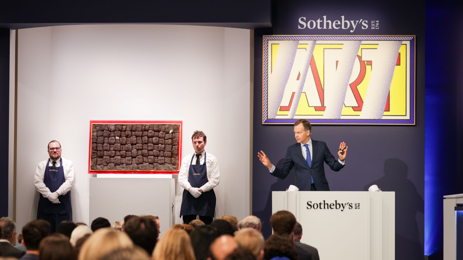 Un banditore d'asta è in piedi su un podio con la scritta "Sotheby's" insieme a due assistenti accanto a un'opera d'arte incorniciata, mentre una folla osserva. Un grande cartello con la scritta "ART" è esposto sulla parete alle loro spalle.