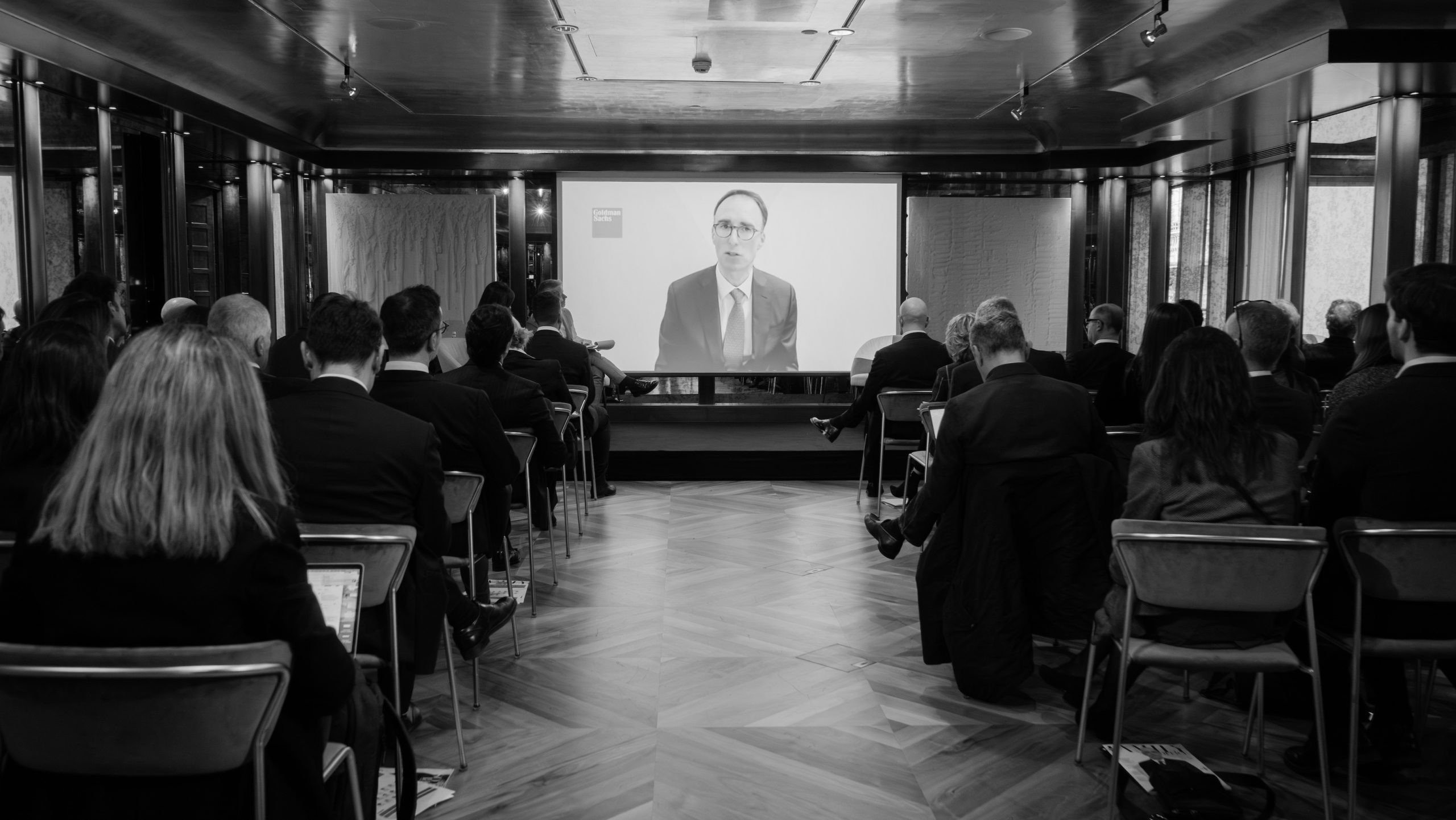 Un gruppo di persone in abiti formali è seduto in fila davanti a un grande schermo, mentre guarda un uomo parlare di infrastrutture in una moderna sala conferenze con pavimenti in legno. L'immagine è in bianco e nero.