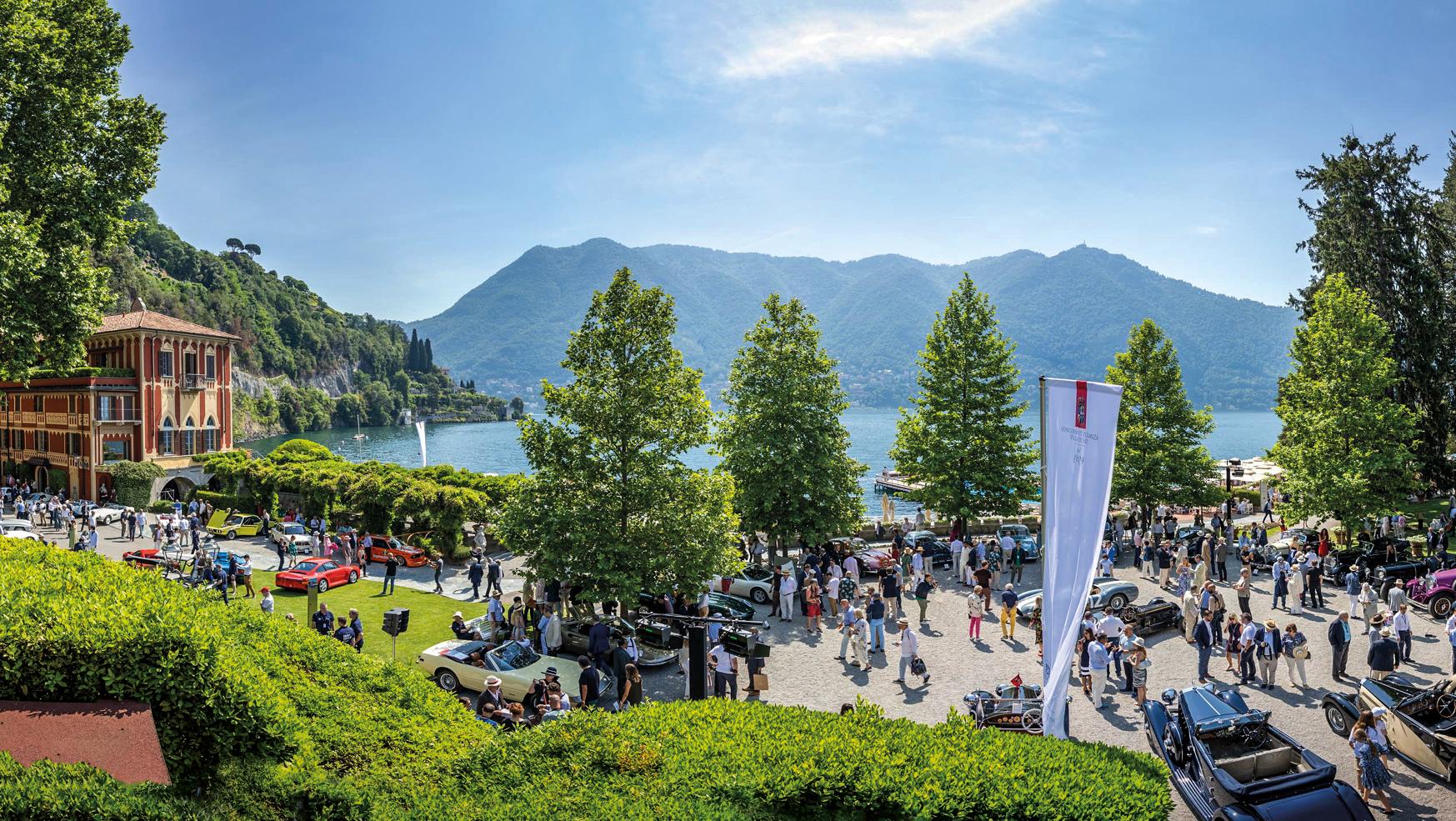 Una vivace mostra automobilistica all'aperto sulle rive di un lago, con auto d'epoca esposte tra gli alberi, una grande folla di persone, un imponente edificio sulla sinistra e montagne sullo sfondo sotto un cielo azzurro e terso.