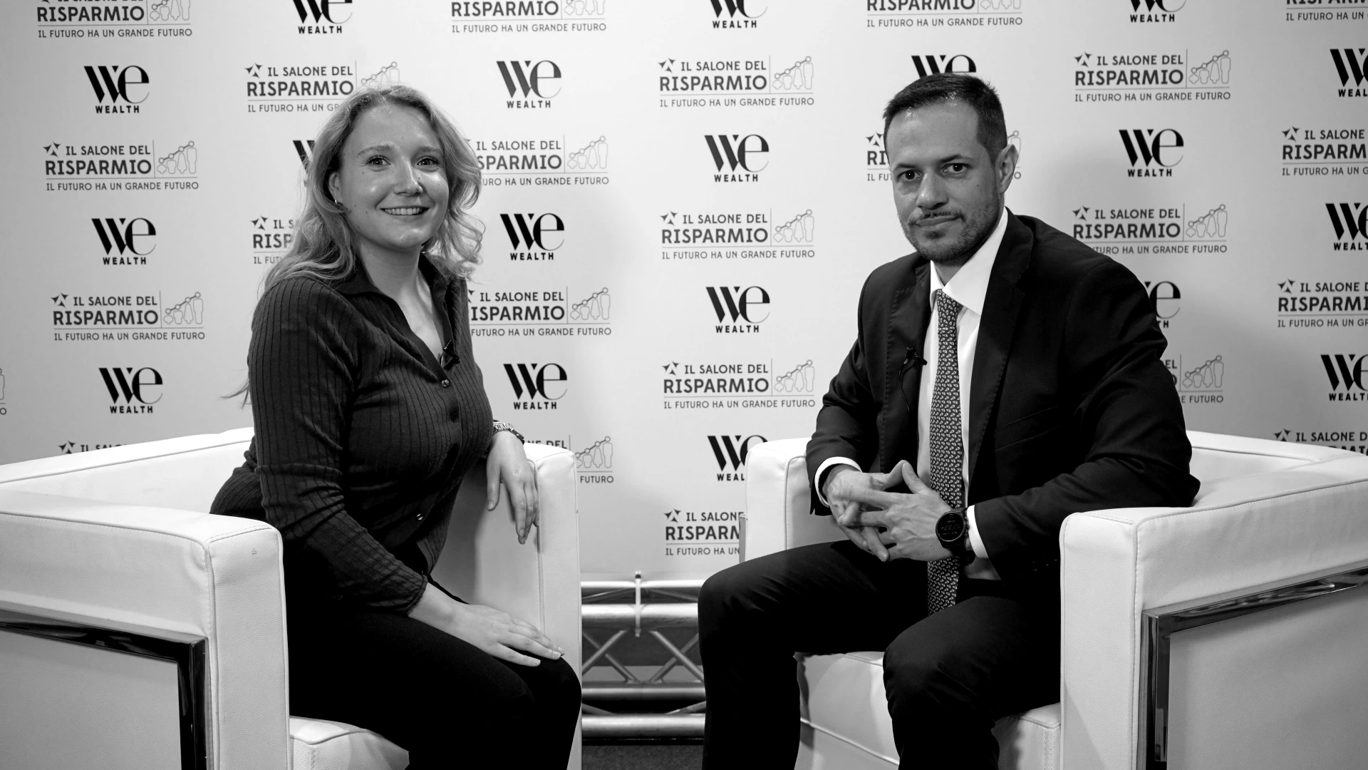 Una donna e un uomo in abito formale siedono su poltrone bianche, uno di fronte all'altro e sorridenti, con uno sfondo decorato con i loghi dell'evento e testi in italiano. L'ambientazione sembra quella di un'intervista o di un dibattito.