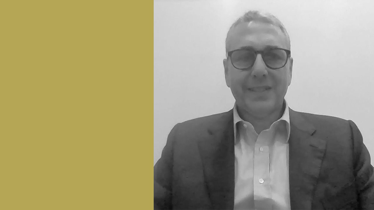 Un uomo con occhiali e giacca sorride alla telecamera, seduto su uno sfondo semplice. La metà sinistra dell'immagine è caratterizzata da un solido blocco di colore giallo senape.