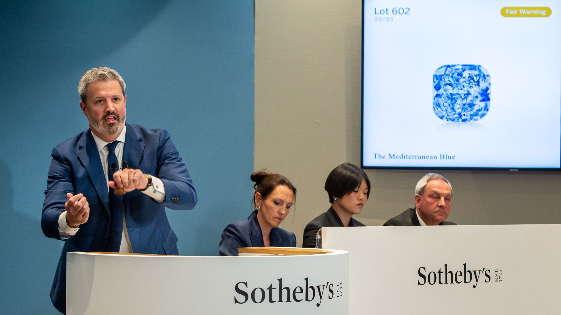 Un banditore di Sotheby's fa un gesto mentre tre membri dello staff siedono dietro di lui. Uno schermo mostra l'immagine del diamante prezioso – un diamante blu con l'etichetta "Lotto 602 - Il Blu Mediterraneo" – con un avviso di "Fair Warning".