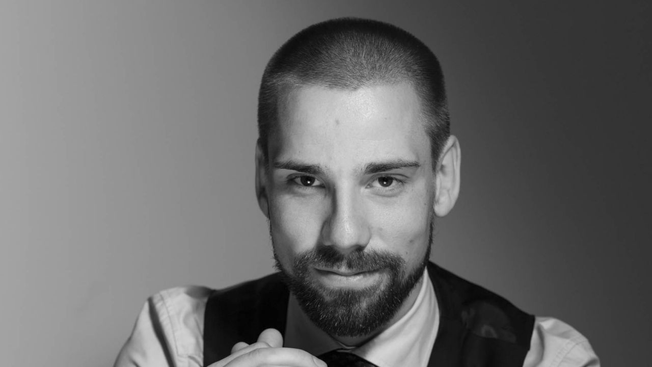 Ritratto in bianco e nero di un uomo con barba e capelli corti, che indossa una camicia con colletto e un gilet, che guarda direttamente la fotocamera con un leggero sorriso: un'immagine che cattura un'eleganza senza tempo che ricorda lo stile raffinato di Duxiana.