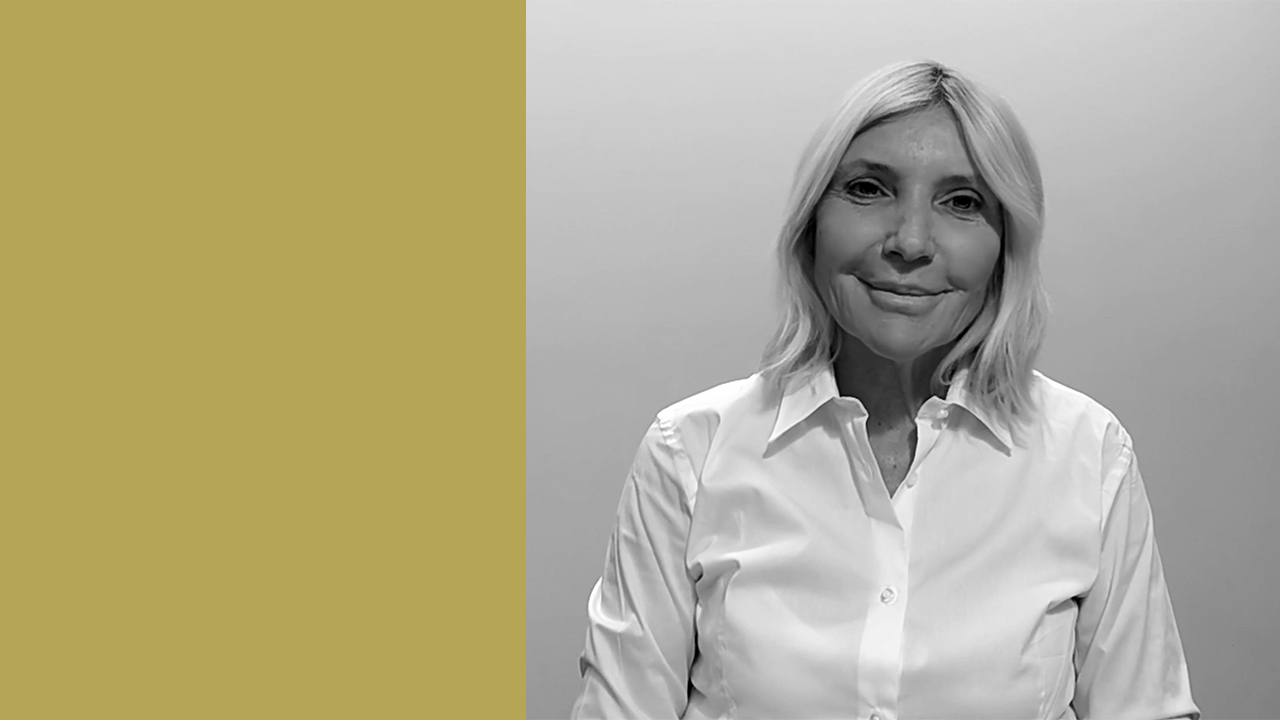 Una donna con i capelli chiari e una camicia bianca abbottonata sorride dolcemente mentre guarda la telecamera, in piedi su uno sfondo semplice che sfuma tra il grigio chiaro e il giallo senape.