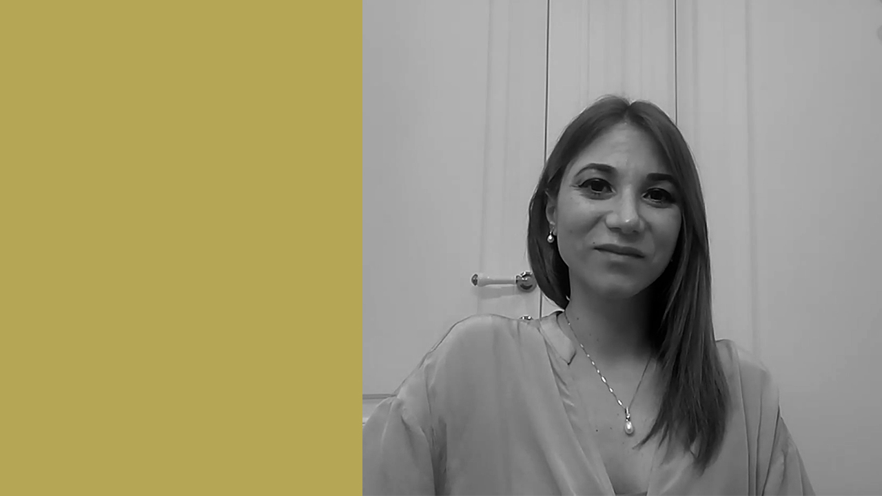 Una donna con i capelli lisci lunghi fino alle spalle e una collana con pendente è seduta in un interno davanti a mobili chiari, sorridendo dolcemente. Il terzo sinistro dell'immagine è di un giallo senape uniforme. L'immagine è in scala di grigi, fatta eccezione per il riquadro giallo.