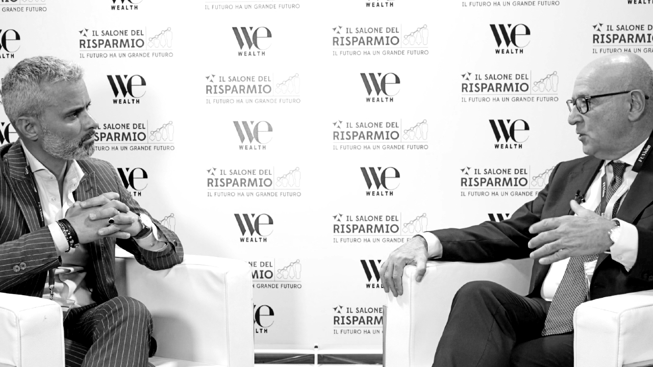 Due uomini in giacca e cravatta, tra cui Manfrone, siedono e conversano animatamente durante un'intervista o un dibattito. Sullo sfondo si vedono i loghi di "WE Wealth" e "Il Salone del Risparmio".