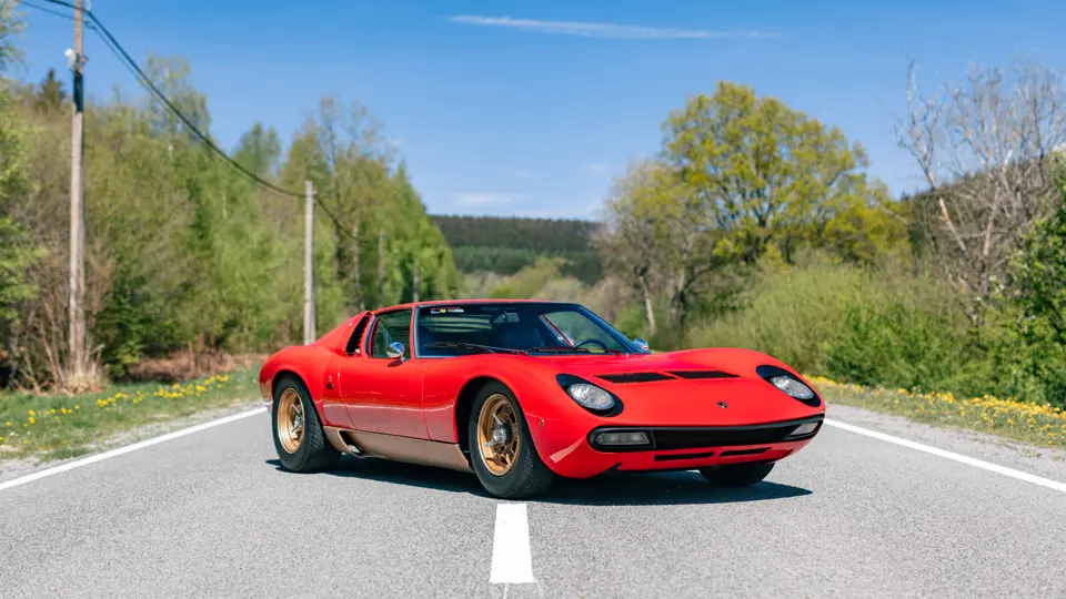 Una classica auto sportiva rossa con dettagli dorati è parcheggiata su una strada deserta e soleggiata, circondata da alberi verdi e cielo azzurro.