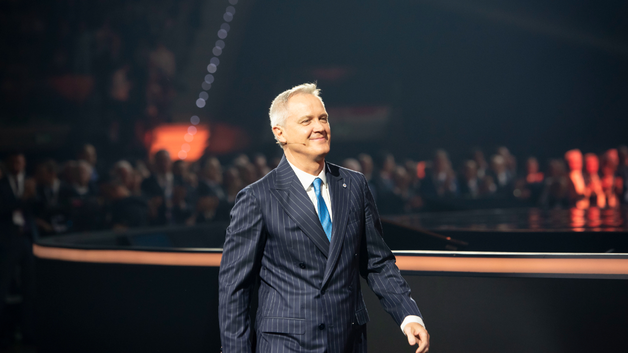 Un uomo in abito gessato e cravatta blu cammina con sicurezza sul palco durante un evento formale di Mediolanum, con il pubblico seduto sullo sfondo sotto un'illuminazione spettacolare.