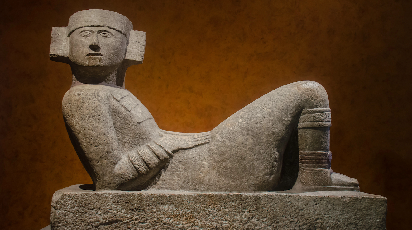Scultura in pietra raffigurante una figura sdraiata con le gambe piegate, un elaborato copricapo e un oggetto appoggiato sul ventre, su uno sfondo marrone.