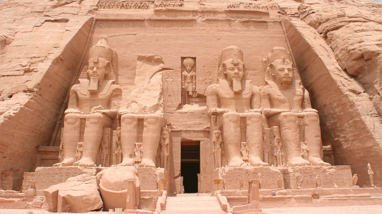 L'immagine mostra la facciata del tempio di Abu Simbel in Egitto, con quattro grandi statue sedute del faraone Ramses II scolpite in una scogliera di arenaria, con statue più piccole e iscrizioni attorno all'ingresso.