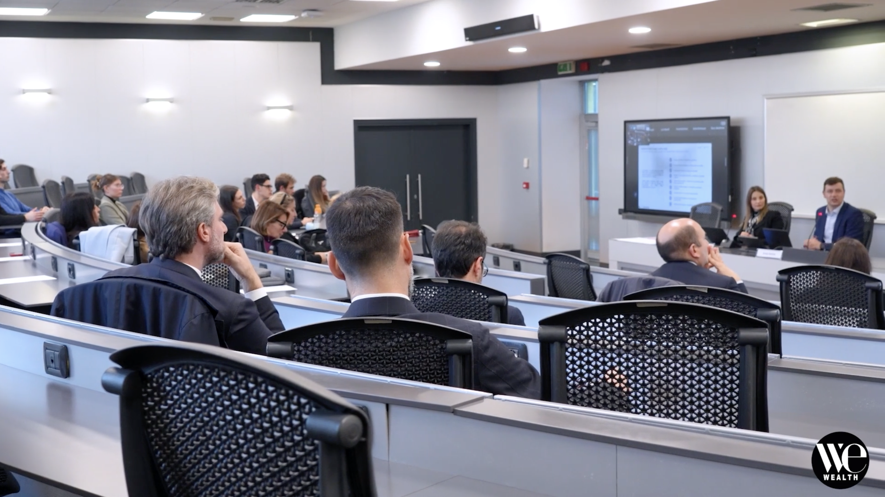 Persone sedute in una moderna aula magna ascoltano una presentazione; due relatori siedono in prima fila, vicino a un grande schermo che mostra un documento. Il logo di WE Wealth appare nell'angolo in basso a destra.